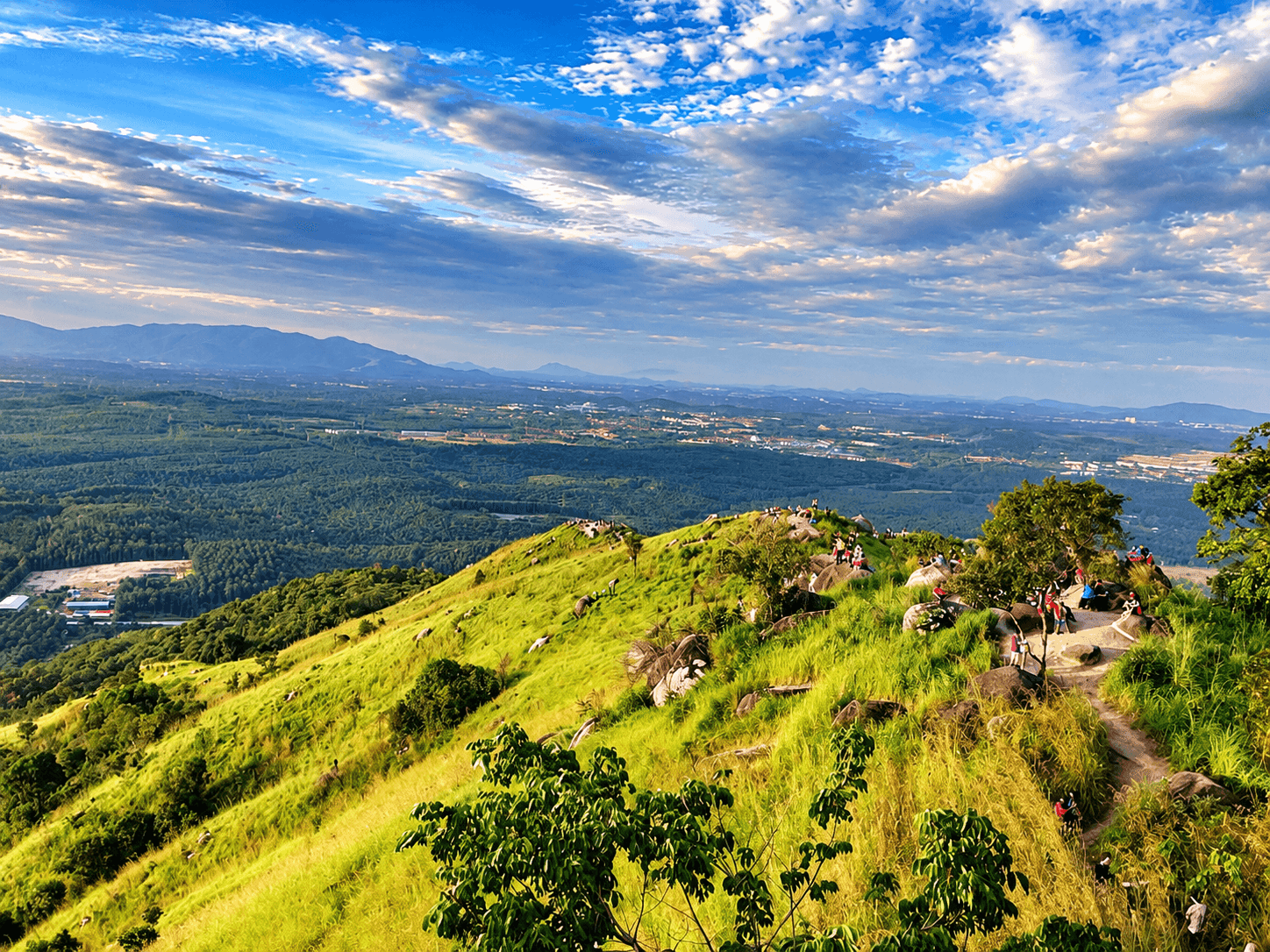 Hikers on the grassy summit of Broga Hill near Semenyih, Selangor, looking out over forest and plantation land at sunrise